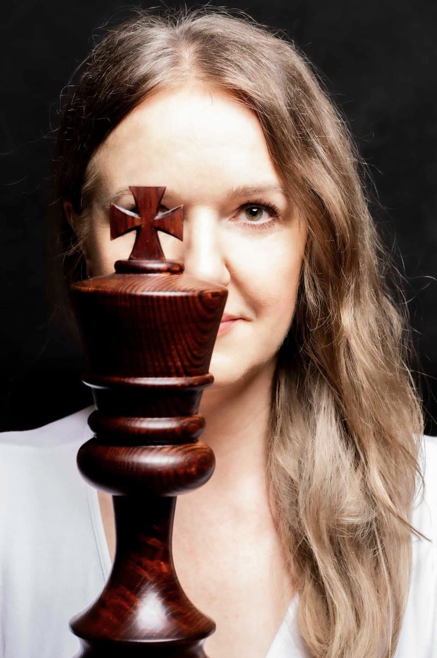 Portret kobiety z długimi włosami, połowę twarzy zasłania drewniana figura szachowego króla